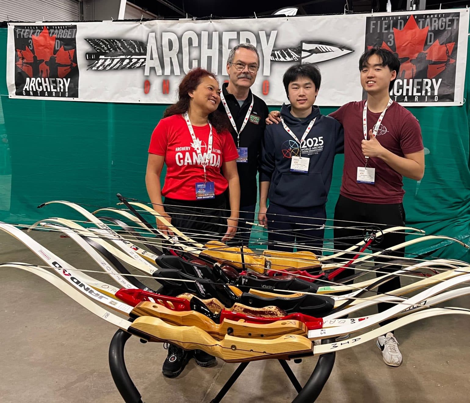 Richard and Lukas with two members of the Archery Ontario booth team, standing behind a rack of recurve bows at the Toronto Sportsmen’s Show.