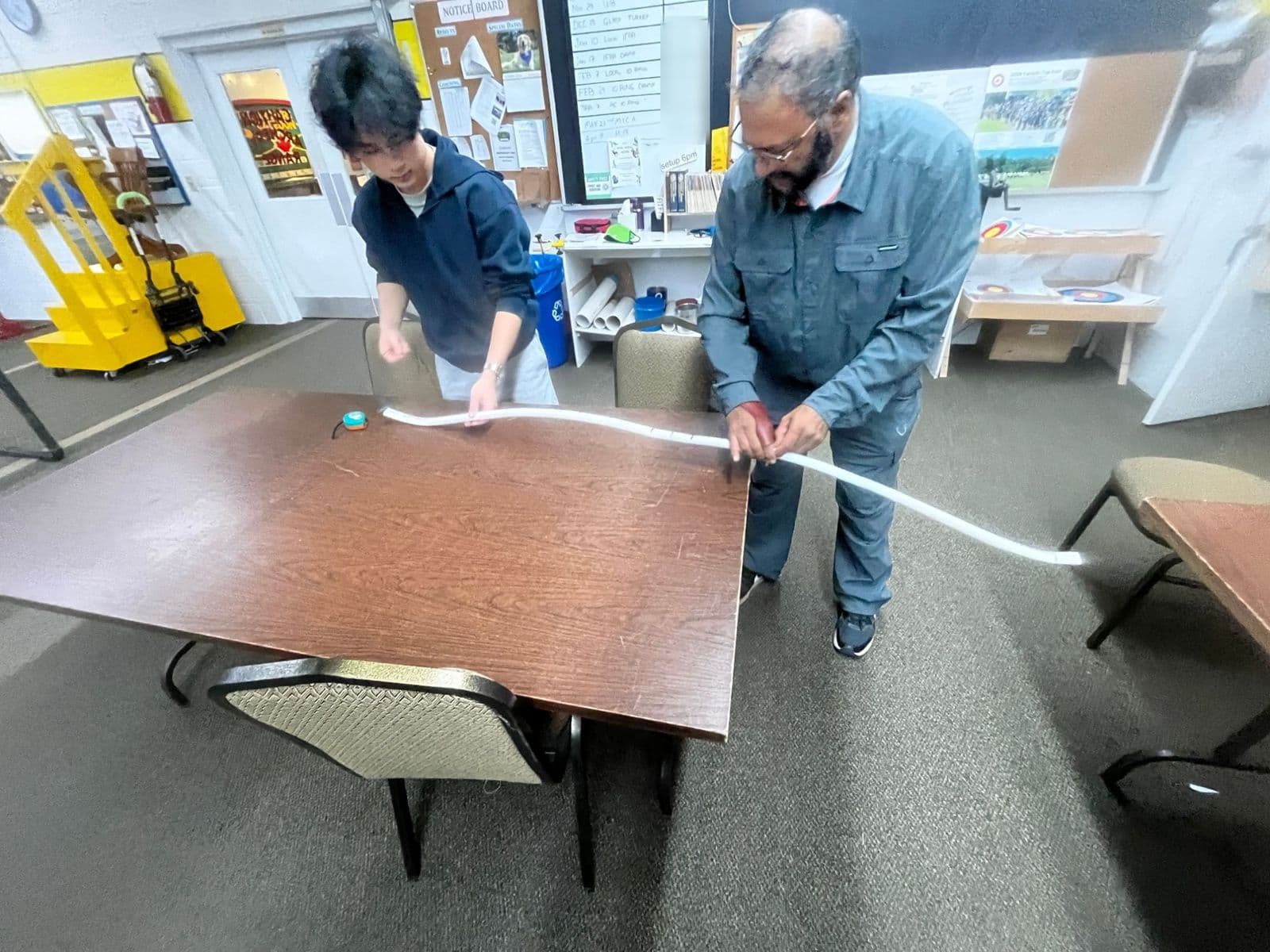 Two people holding a bent PVC bow across a table in the clubhouse to check its curve.