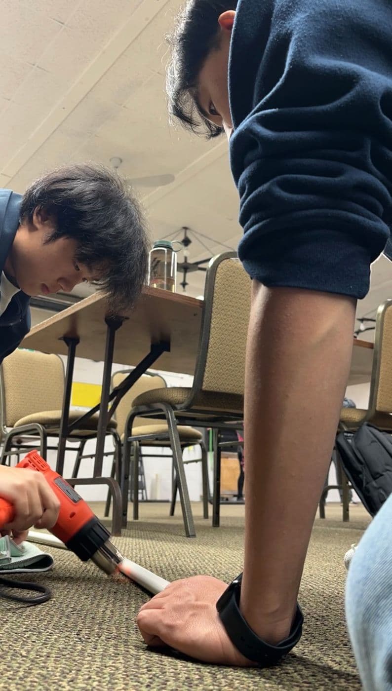 Two founders on the floor, one warming the end of a PVC pipe with a heat gun while the other holds it down.